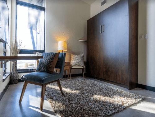 Cozy room featuring a chair, a small table, a lamp, and a wall bed, with a textured rug on the floor.