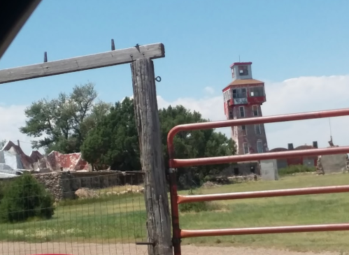 A tall, red and white tower stands near a rustic gate, with green trees and a blue sky in the background.