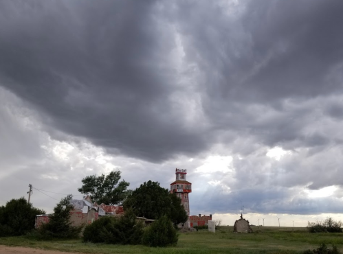 A tall, weathered tower stands amidst dark, stormy clouds and green vegetation in a rural landscape.