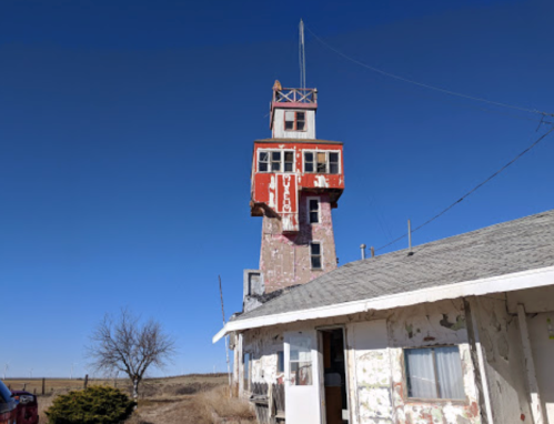 A weathered tower with a red and white structure stands beside an old building under a clear blue sky.