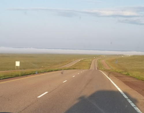 A wide, open highway stretches into the distance, flanked by grassy fields under a clear sky.