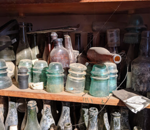 A collection of dusty glass bottles and jars on a wooden shelf, showcasing various shapes and sizes.