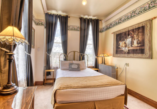 Cozy bedroom with a bed, decorative lamps, curtains, and artwork on the walls, featuring warm tones and natural light.