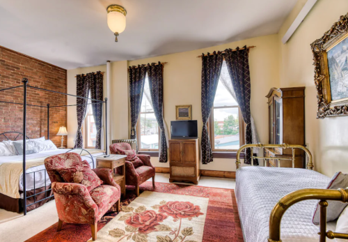 Cozy bedroom with a vintage aesthetic, featuring a bed, armchairs, and large windows with patterned curtains.