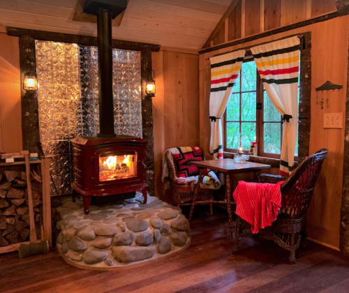 Cozy cabin interior with a wood stove, stone floor, and rustic decor, featuring a table and chairs by a window.