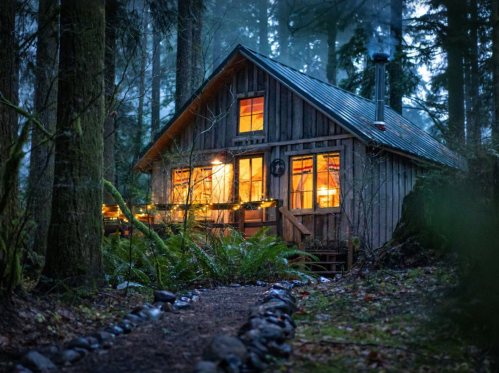 A cozy wooden cabin illuminated at dusk, surrounded by tall trees and a misty forest.