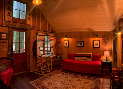 Cozy cabin interior with wooden walls, a red couch, a patterned rug, and snowshoes decor. Natural light from windows.