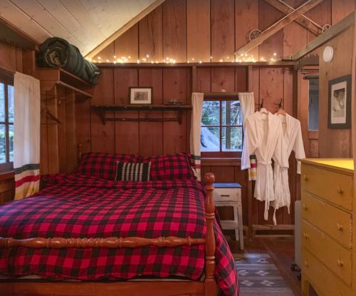 Cozy cabin interior with a wooden bed, plaid bedding, yellow dresser, and warm lighting. Robes hang by the window.