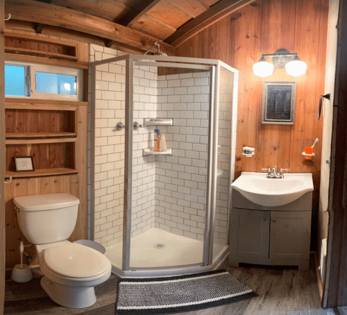 A cozy bathroom featuring a shower, toilet, sink, wooden walls, and a small shelf.
