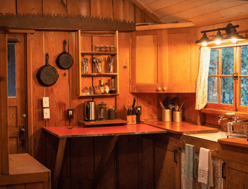 Cozy wooden kitchen with a red countertop, hanging pots, and warm lighting, featuring a window and organized shelves.