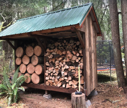 A wooden shed with a green metal roof, stacked with firewood on one side, surrounded by trees.