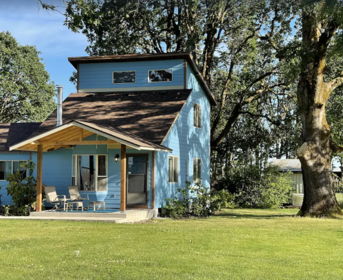 A blue two-story house with a porch, surrounded by green grass and trees under a clear blue sky.