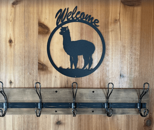 A wooden wall with a black llama silhouette and "Welcome" sign above a row of six metal hooks.