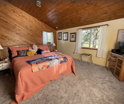 Cozy bedroom with a wooden ceiling, a large bed, and a window overlooking greenery. Soft carpet and decorative elements.