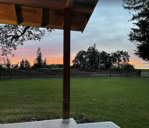 A serene sunset view from a porch, with colorful skies and a grassy yard framed by trees and a wooden fence.