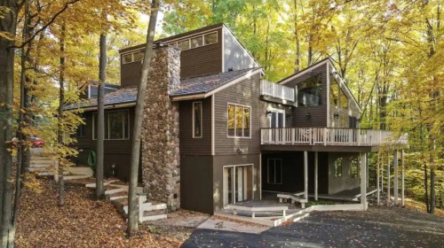 A modern house surrounded by autumn trees, featuring a stone chimney and multiple levels with a deck.