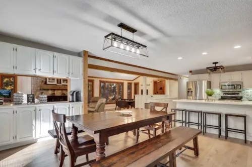 Bright, modern kitchen and dining area with wooden table, white cabinets, and large windows showcasing natural light.
