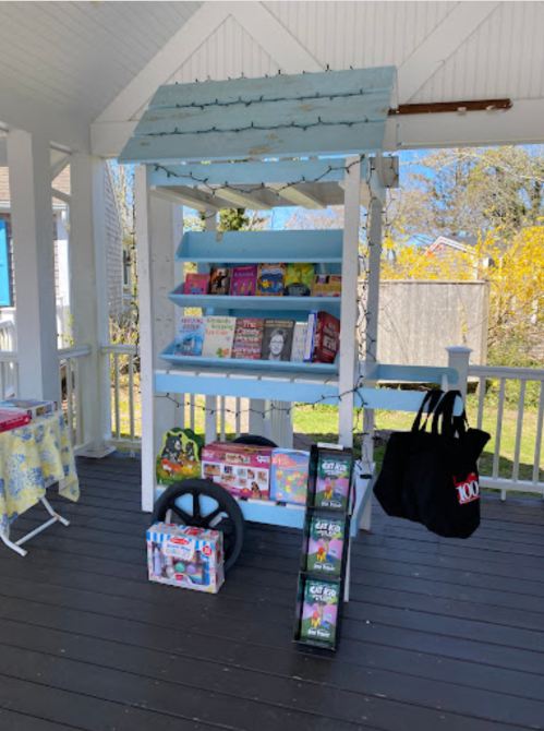 A wooden book stand with shelves displaying various books, a tote bag, and a wheel, set in a sunny outdoor area.