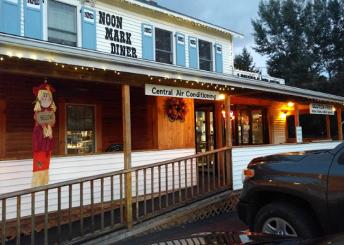 Exterior of Noon Mark Diner with a welcoming sign, lights, and a scarecrow decoration. Evening setting.