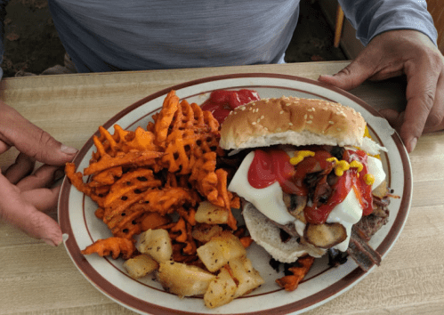 A plate with a burger topped with mayonnaise, ketchup, and mustard, alongside sweet potato fries and seasoned potatoes.
