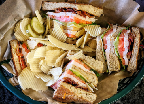 A basket containing a club sandwich with bacon, lettuce, and tomato, accompanied by a side of potato chips and pickles.