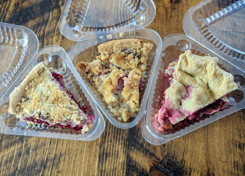 Three slices of pie in clear plastic containers, featuring crumbly toppings and fruit fillings, arranged on a wooden surface.