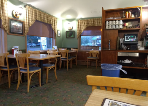 Cozy diner interior with wooden tables, chairs, and a counter, featuring green walls and large windows with checkered curtains.