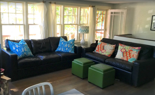 A cozy living room with two black sofas, colorful patterned pillows, green ottomans, and large windows letting in natural light.