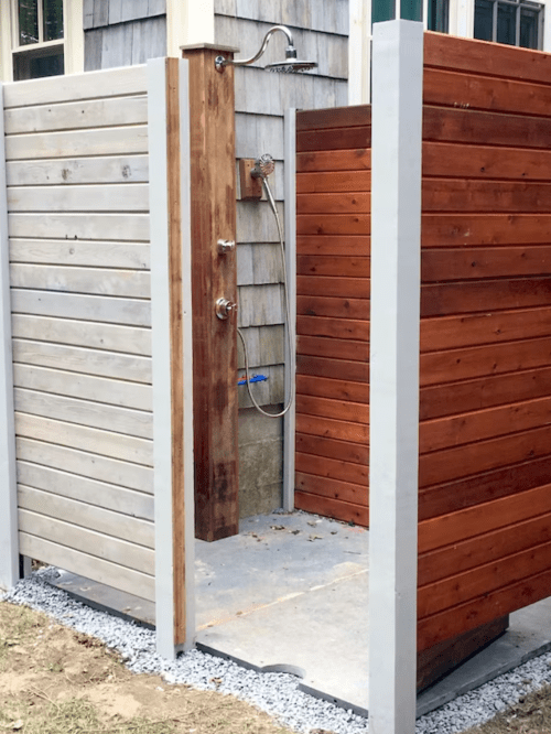 An outdoor shower enclosed by wooden and gray paneling, featuring a showerhead and stone flooring.