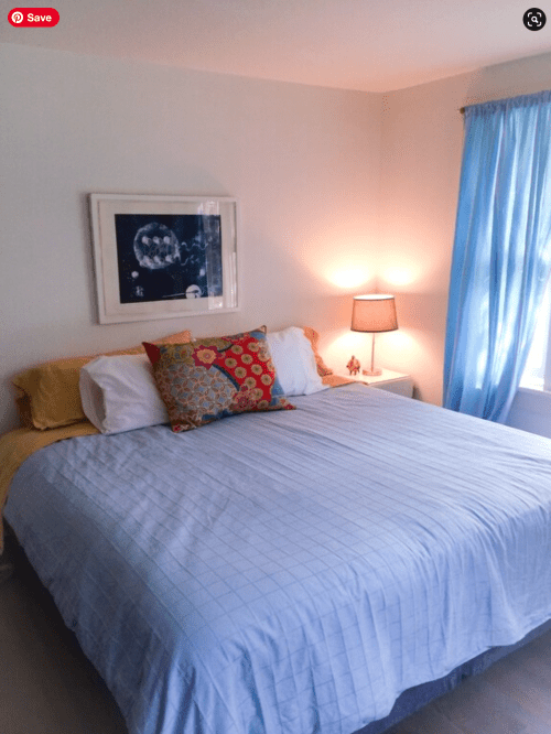 Cozy bedroom with a large bed, colorful pillow, lamp, and blue curtains, featuring a framed artwork on the wall.