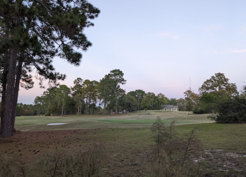A serene landscape featuring a golf course surrounded by trees under a pastel sky at dusk.