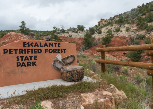 Sign for Escalante Petrified Forest State Park, with a rocky landscape and wooden fence in the background.