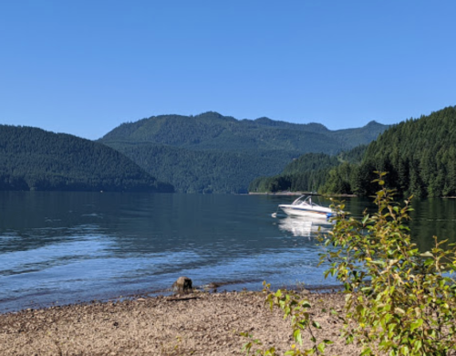 A serene lake scene with a boat on the water, surrounded by mountains and lush greenery under a clear blue sky.
