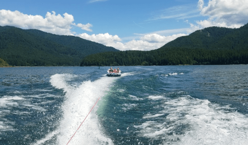 A boat pulls people on a tube across a lake, surrounded by mountains and blue skies with fluffy clouds.