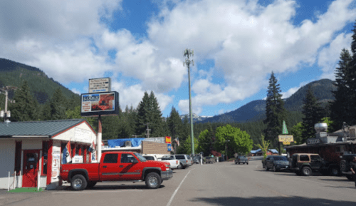 A small mountain town street with shops, parked cars, and trees under a partly cloudy sky.