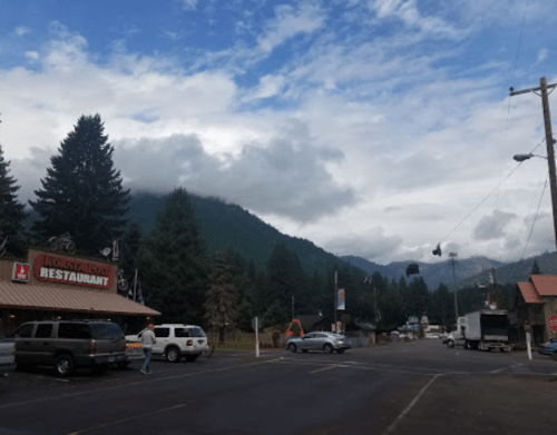 A small mountain town with a restaurant, parked cars, and trees under a cloudy sky. Mountains are visible in the background.