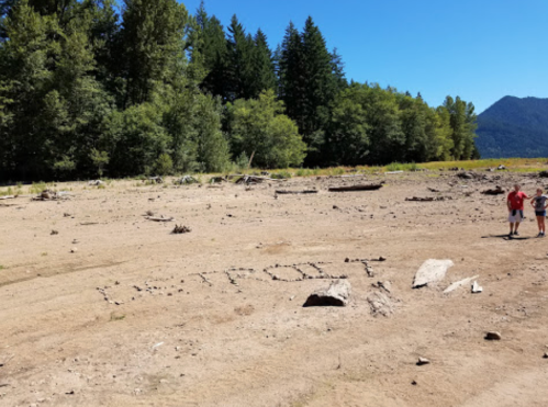 Two people stand on a dry, cracked riverbed with "DETROIT" written in the sand, surrounded by trees and mountains.