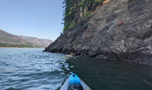 A kayak on calm water near a rocky shoreline, surrounded by trees and mountains under a clear blue sky.