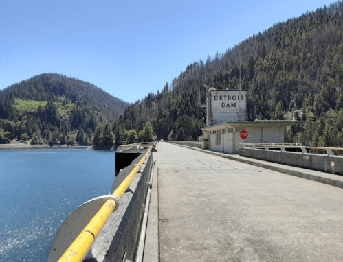 A view of Detroit Dam with a stop sign, surrounded by mountains and a calm blue lake under a clear sky.