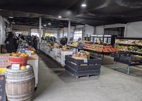 A bustling indoor market with various fruits and vegetables displayed on tables and crates, and shoppers browsing.