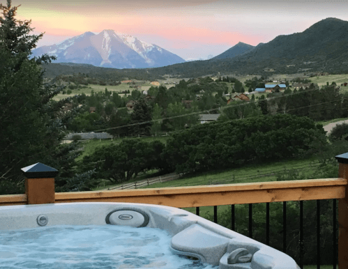 A hot tub on a deck overlooks a scenic landscape with mountains and a sunset in the background.