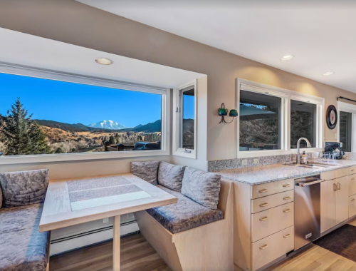 Bright kitchen with a cozy dining nook and large windows showcasing a mountain view. Modern appliances and light decor.