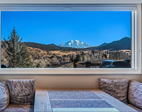 A cozy interior view through a large window showcasing a mountain landscape under a clear blue sky.