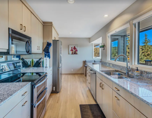 Modern kitchen with light wood cabinets, granite countertops, stainless steel appliances, and large windows.