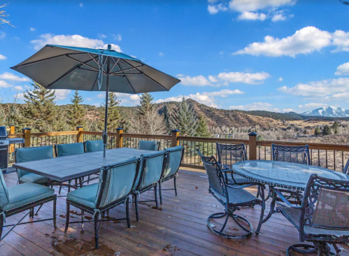 Outdoor deck with seating, umbrella, and scenic mountain views under a blue sky with fluffy clouds.