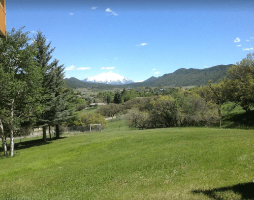 A scenic view of green fields and trees, with snow-capped mountains in the background under a clear blue sky.