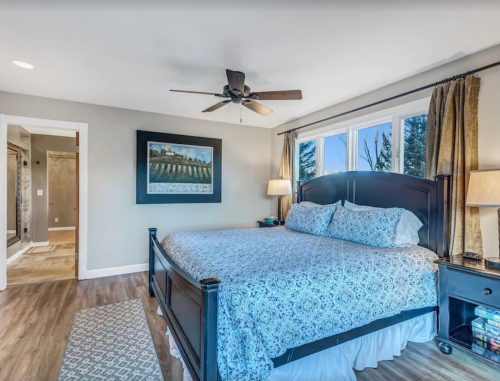 Bright bedroom with a large bed, ceiling fan, and artwork, featuring a connected bathroom and natural light from windows.