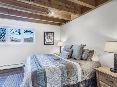 Cozy bedroom with a large bed, wooden beams, and a window showcasing a scenic view. Soft lighting and decor add warmth.