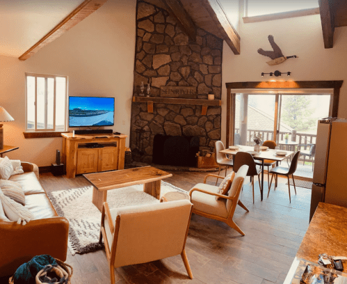 Cozy living room with a stone fireplace, wooden furniture, and large windows overlooking a deck.