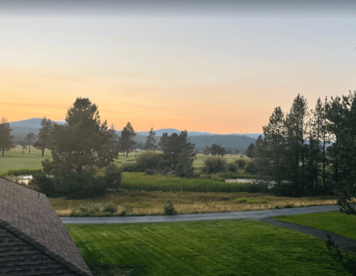 A serene landscape at sunrise, featuring trees, a golf course, and distant mountains under a colorful sky.